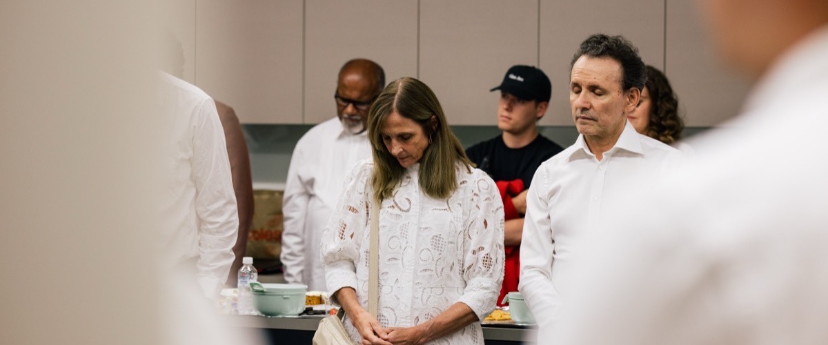 A group of people in white shirts stand in a room with a table, with some individuals facing forward and others turned away.