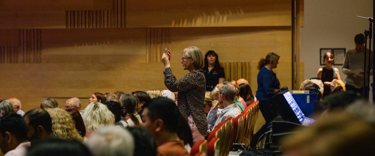 A woman stands in a crowded auditorium, holding a small device in her hand, with an audience seated in front of her.