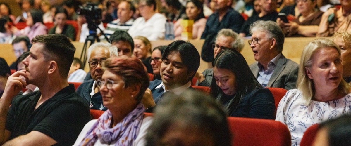 Graph depicts a crowd of people seated in an auditorium, with a camera positioned in front of them.
