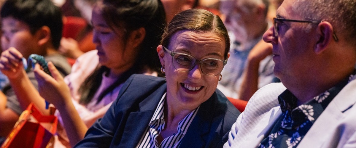 A woman in a navy blazer sits among a crowd of people, wearing glasses and a striped shirt.