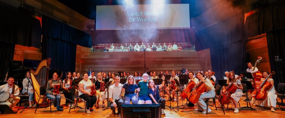 A large group of people posing on a stage with musical instruments, including cellos and harps.