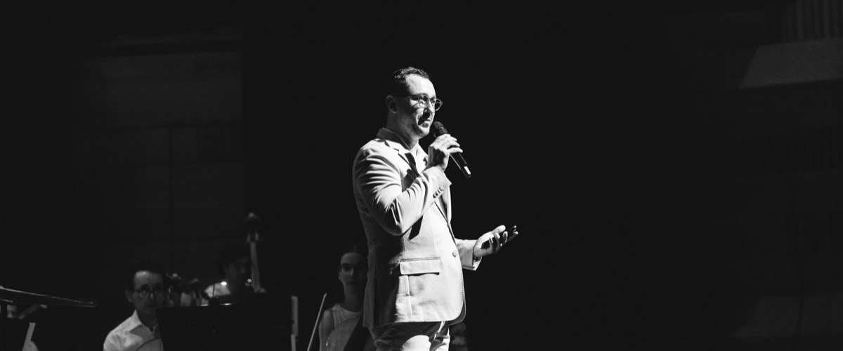 This black-and-white photograph depicts a man in a suit singing into a microphone, accompanied by musicians in the background.