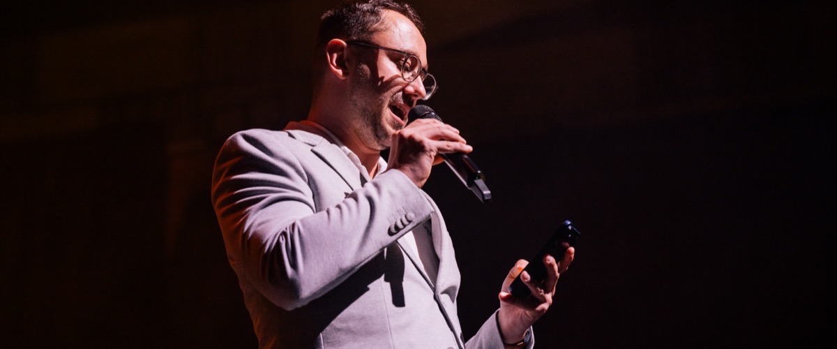 A man in a light-colored suit jacket, holding a microphone and a small black object, possibly a remote control.