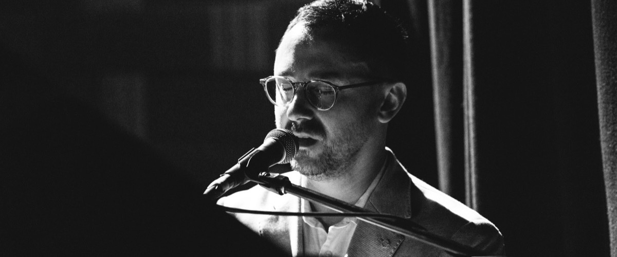 This black-and-white photograph depicts a man singing into a microphone, with a spotlight illuminating his face.
