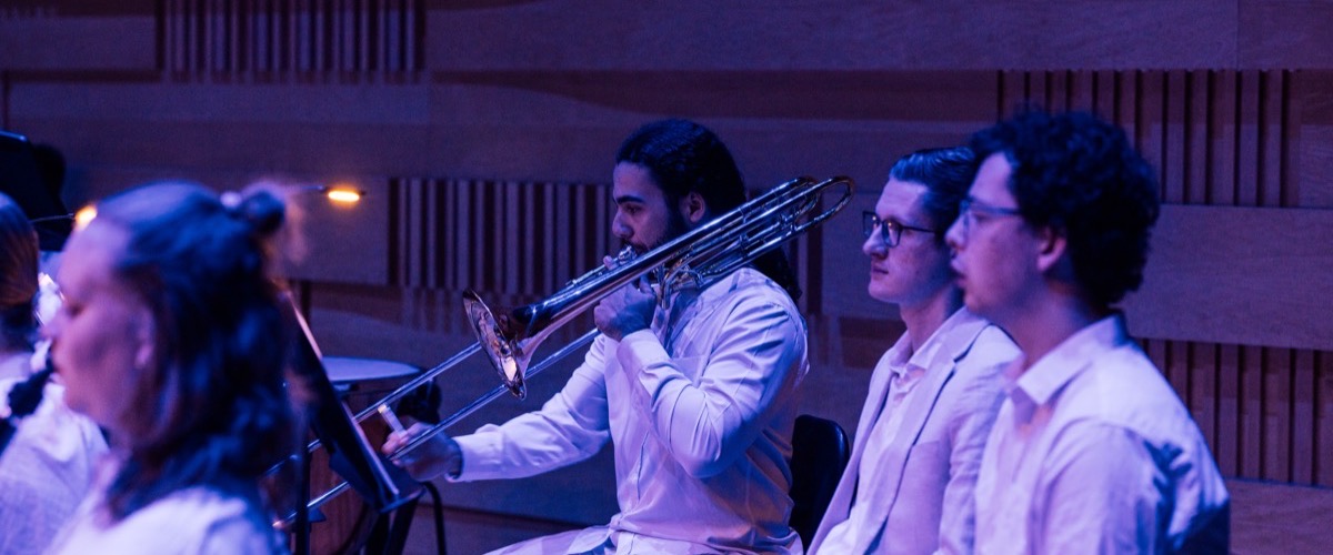 A group of musicians in a dimly lit room, with a man in the center playing a trombone.