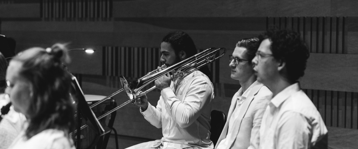 This black-and-white photograph depicts a trombone player seated among other musicians in a dimly lit room.