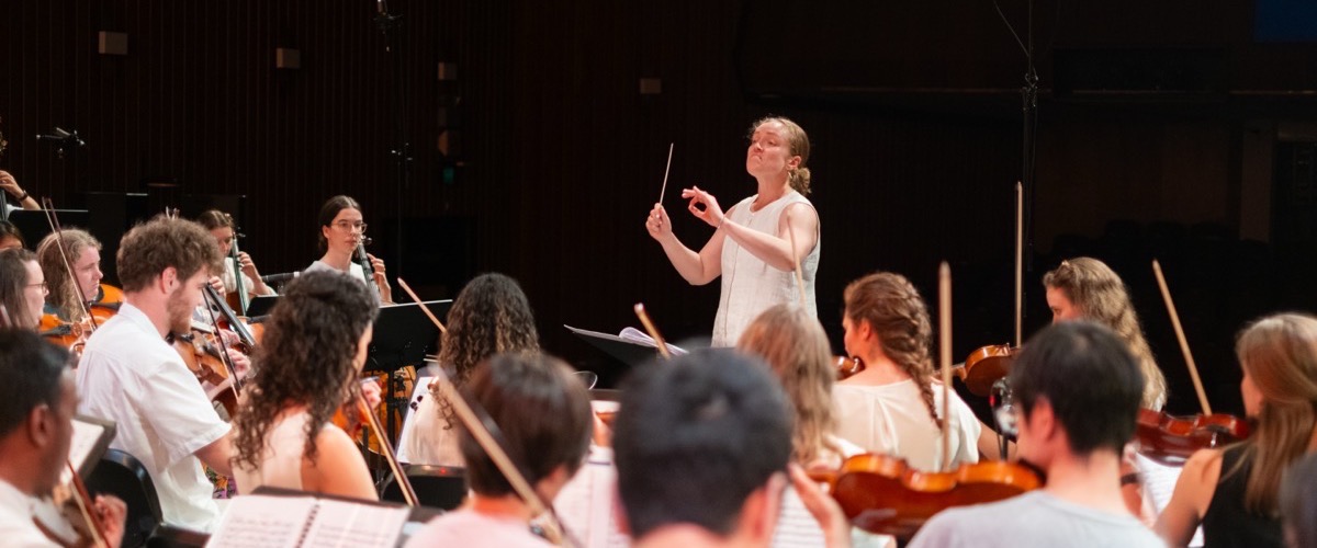 The photograph shows a woman in a white sleeveless dress conducting a large group of people playing stringed instruments.
