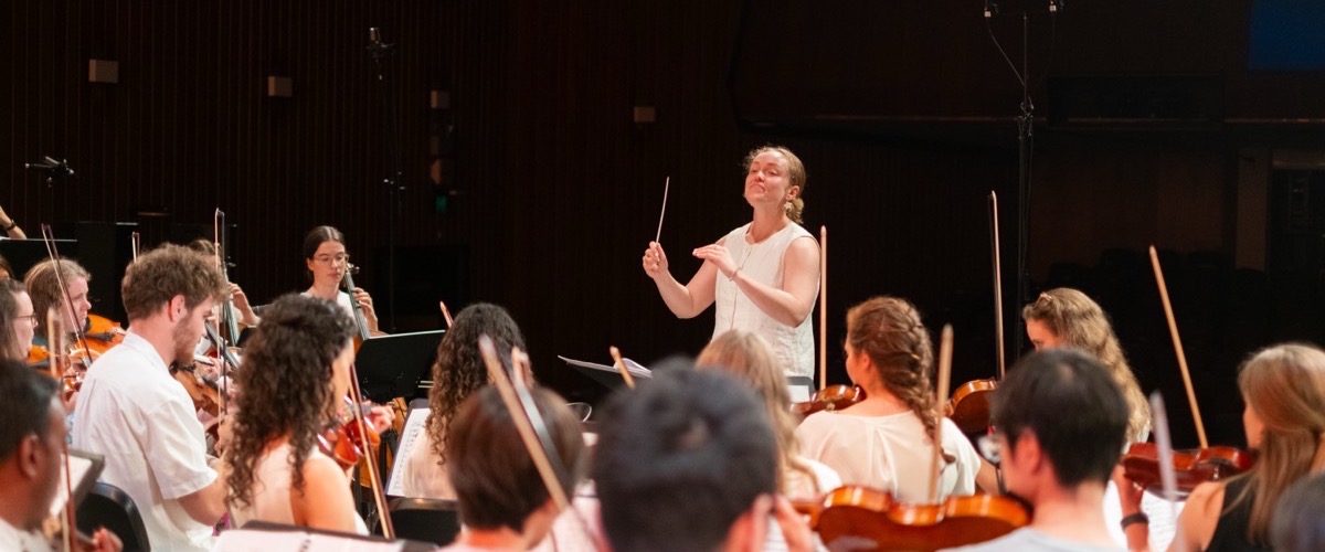 A conductor leads an orchestra, standing at the front with a baton in hand.