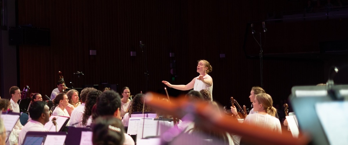 A woman conducting an orchestra, with musicians seated in front of her.
