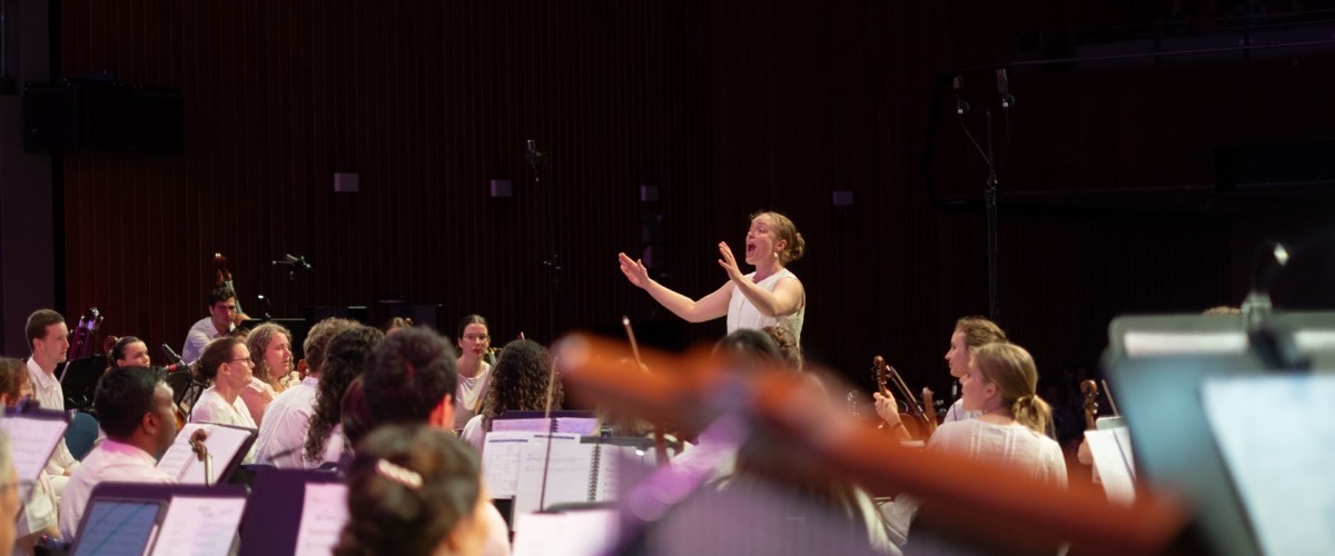 A woman stands in front of a group of musicians, directing them in a performance.