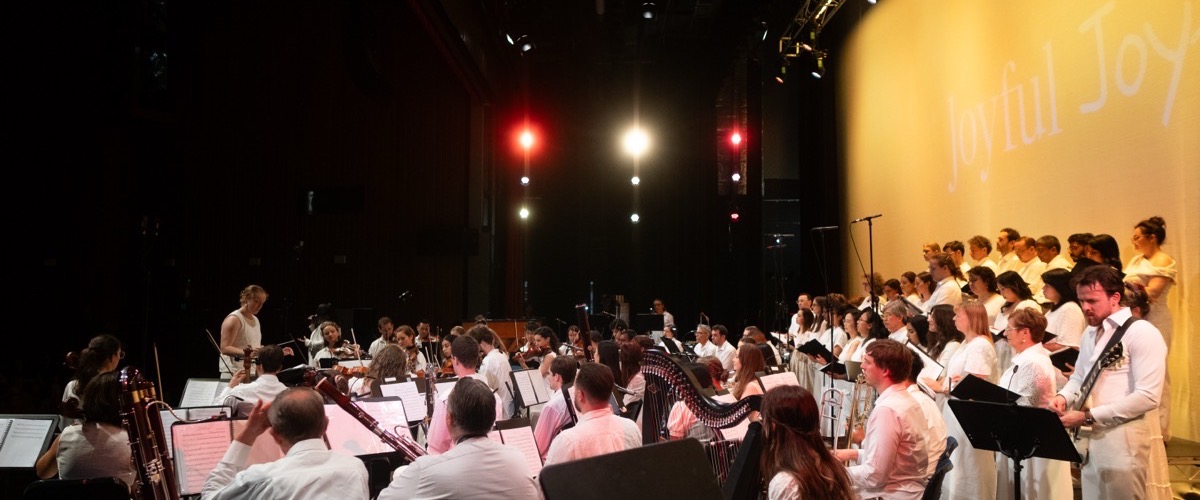 A large group of people, dressed in white, are playing musical instruments on a stage with a large screen displaying the words "Joyful Joy".