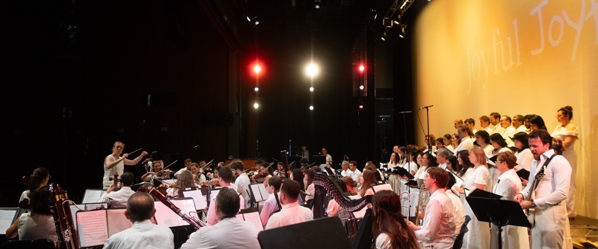 A large ensemble of musicians and singers performing on a stage, with a prominent yellow wall featuring the word "Joyful" in white.