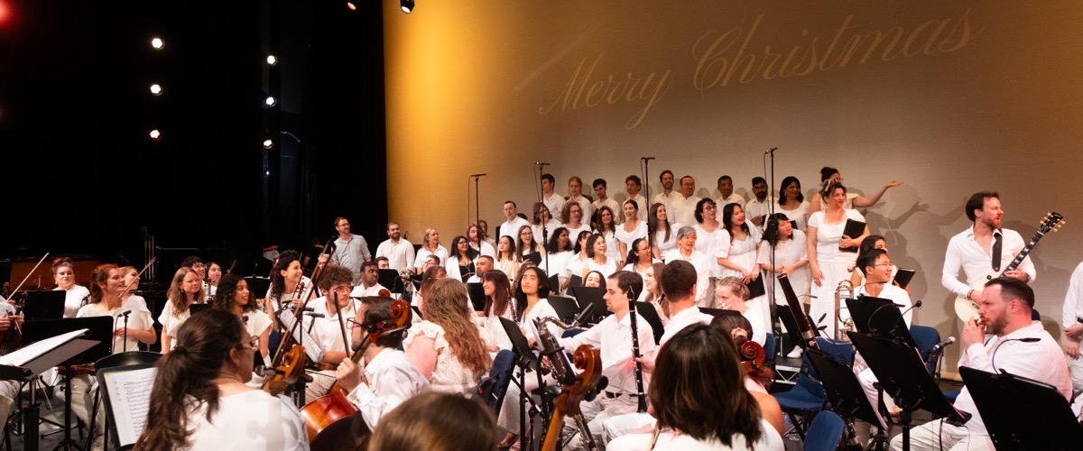 Graph depicts a large group of musicians and singers performing on stage, with a "Merry Christmas" message projected on the wall.